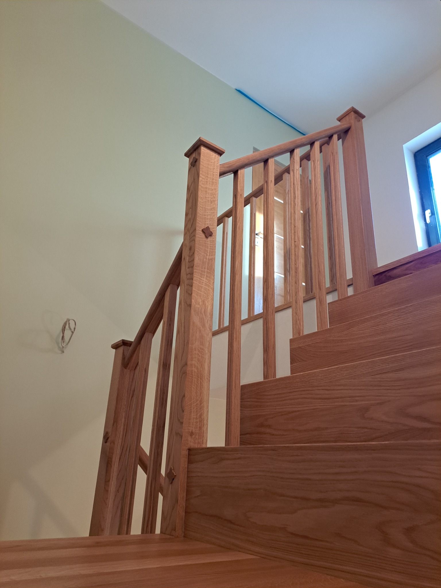 Drewniane schody z jasnego drewna z balustradą, widok z dołu, na ścianie widoczne kable elektryczne.