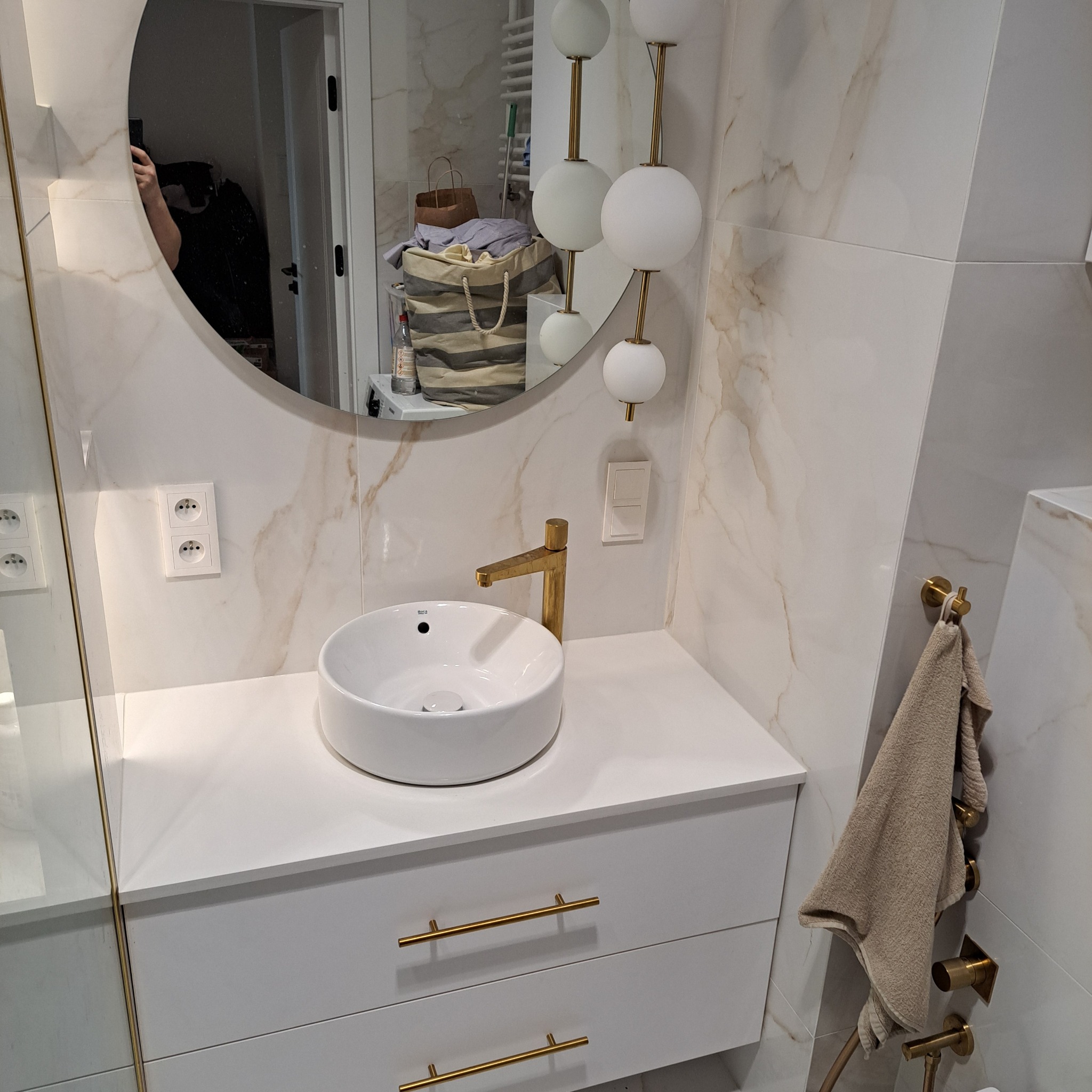 Nowoczesna łazienka z umywalką nablatową, złotą armaturą i okrągłym lustrem. Na ścianie marmurowe płytki i dekoracyjne oświetlenie kulkowe. Widoczny fragment kabiny prysznicowej i ręcznik na złotym...