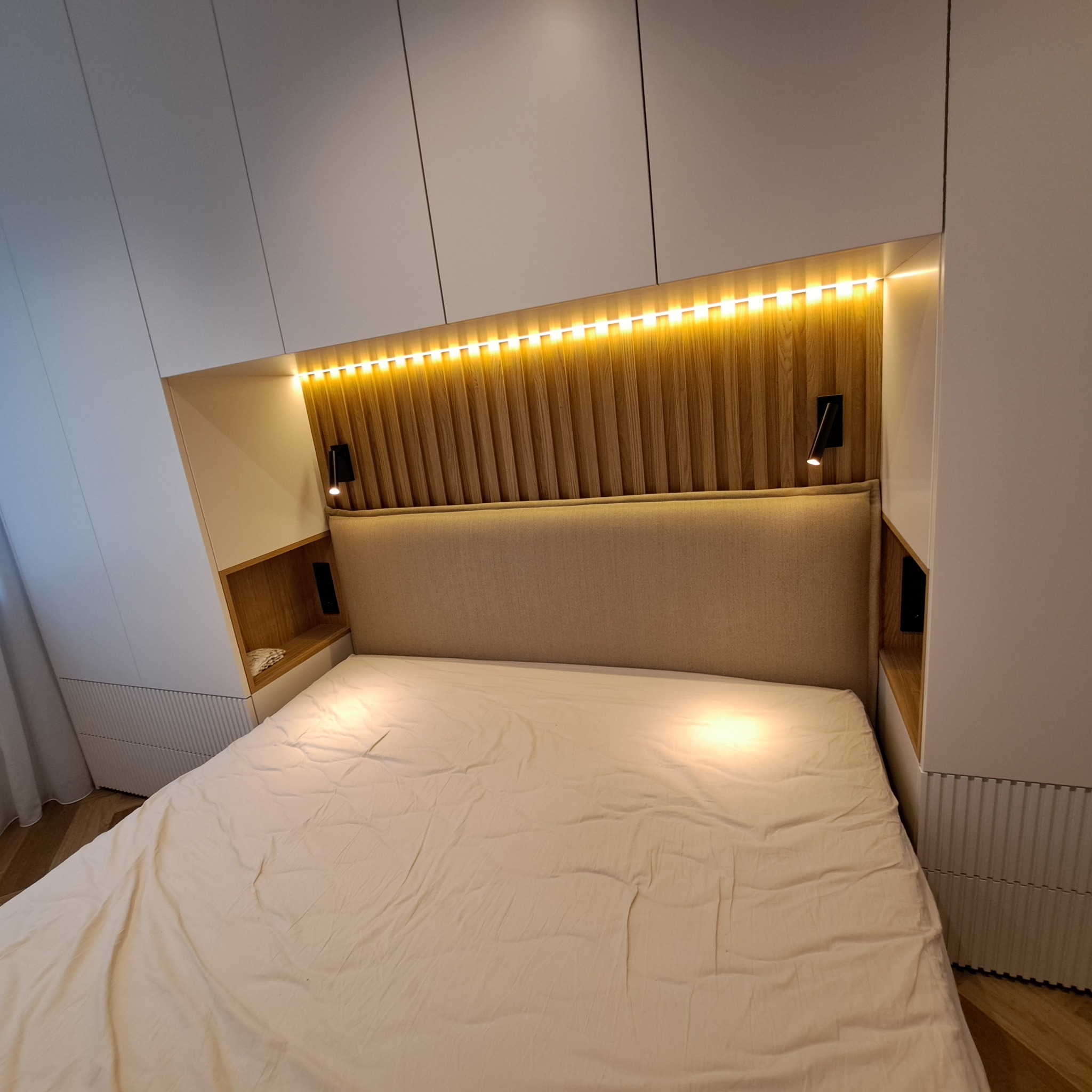 Nowoczesna sypialnia z zabudową meblową: białe szafy, drewniane panele za wezgłowiem łóżka z oświetleniem LED i lampkami nocnymi.