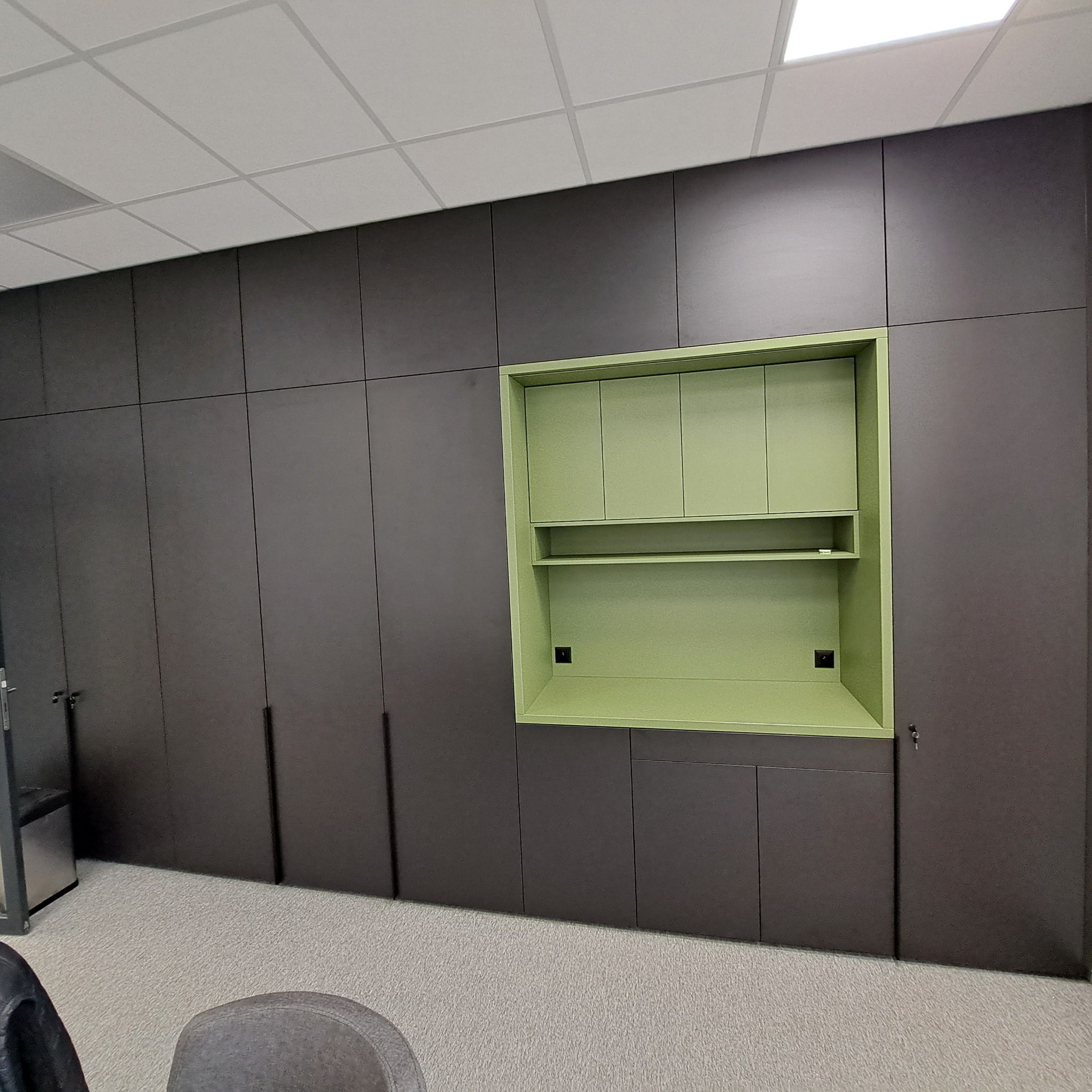 Długa zabudowa meblowa w kolorze grafitowym z wnęką i szafkami w odcieniu pistacjowej zieleni, z widocznymi gniazdkami elektrycznymi we wnęce, na tle jasnej wykładziny dywanowej.