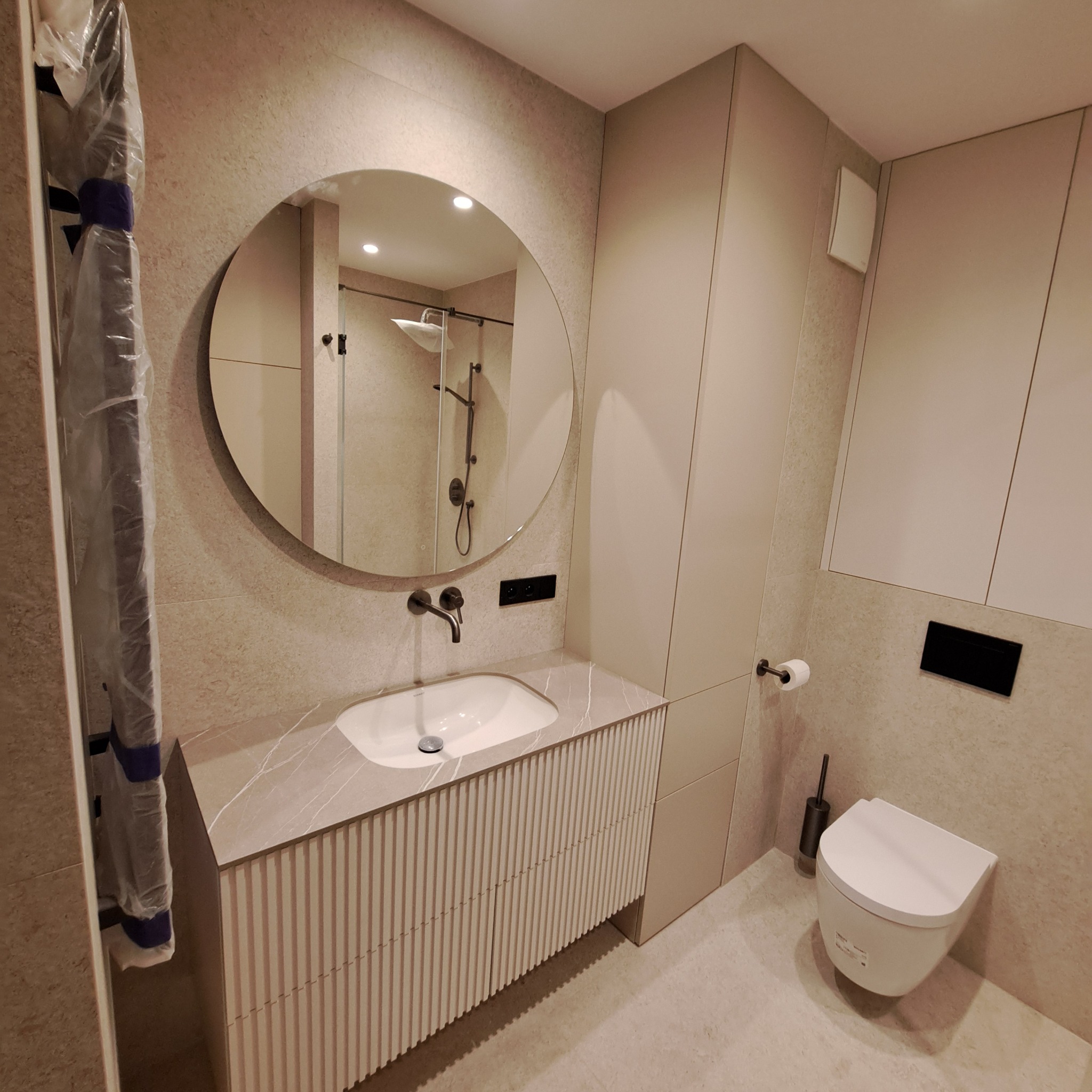 Nowoczesna łazienka w beżach z okrągłym lustrem, umywalką nablatową na szafce z ryflowanym frontem i wiszącą toaletą. Grzejnik drabinkowy w folii ochronnej.