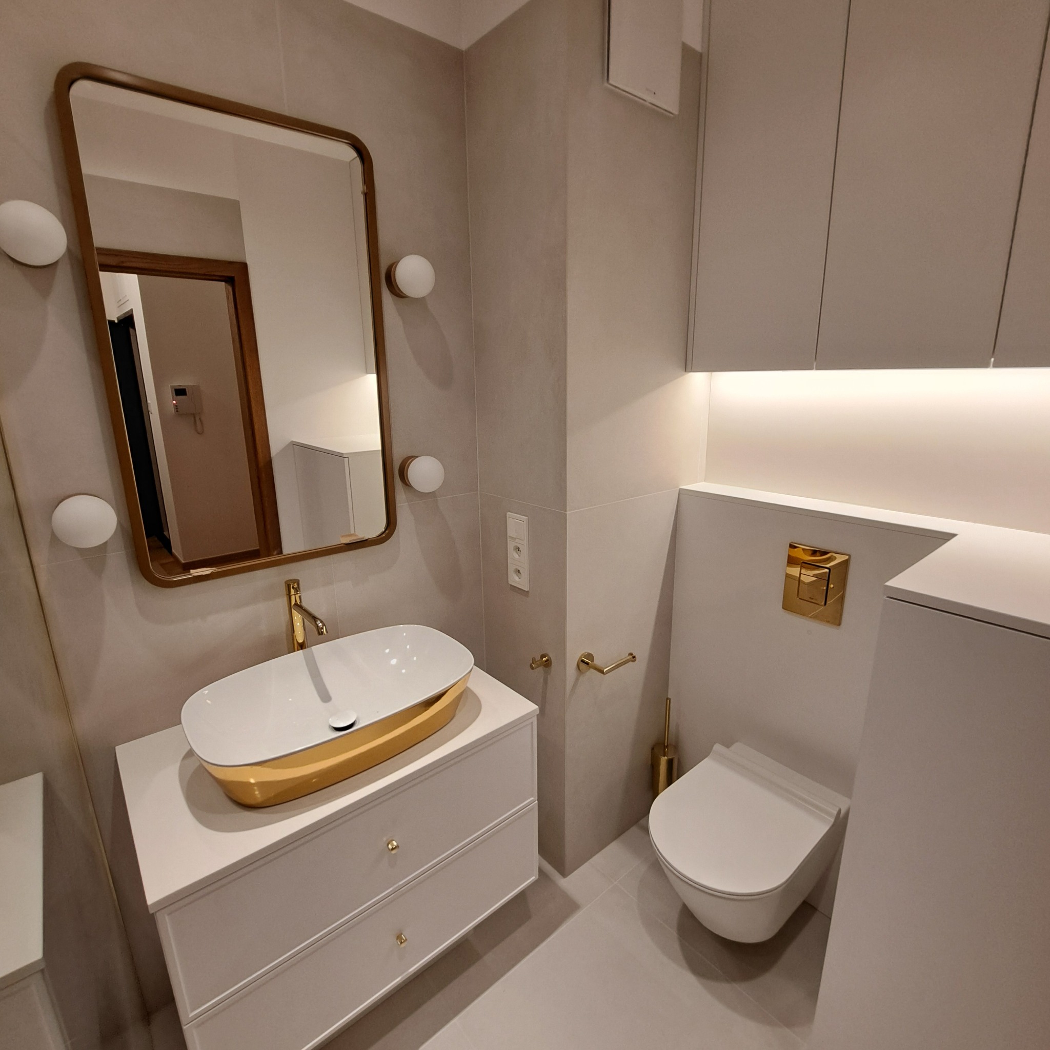 Nowoczesna łazienka w odcieniach szarości z białymi meblami i złotymi akcentami: owalna umywalka na szafce z szufladami, wisząca toaleta z przyciskiem w złotym kolorze, lustro w złotej ramie...