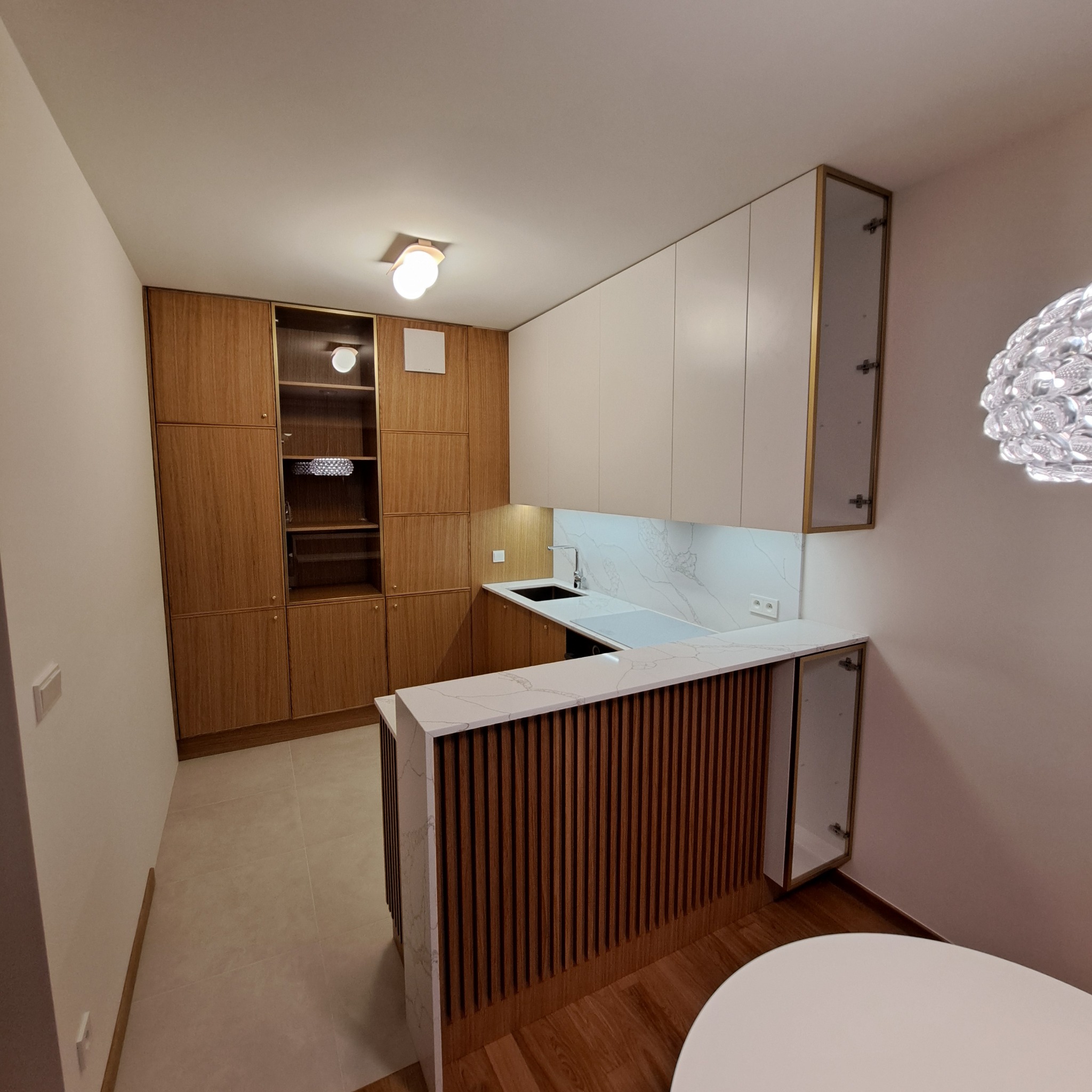 Nowoczesna kuchnia z drewnianymi szafkami, białymi frontami górnych szafek i blatem imitującym marmur, oświetlona lampą sufitową i punktowym światłem pod szafkami.