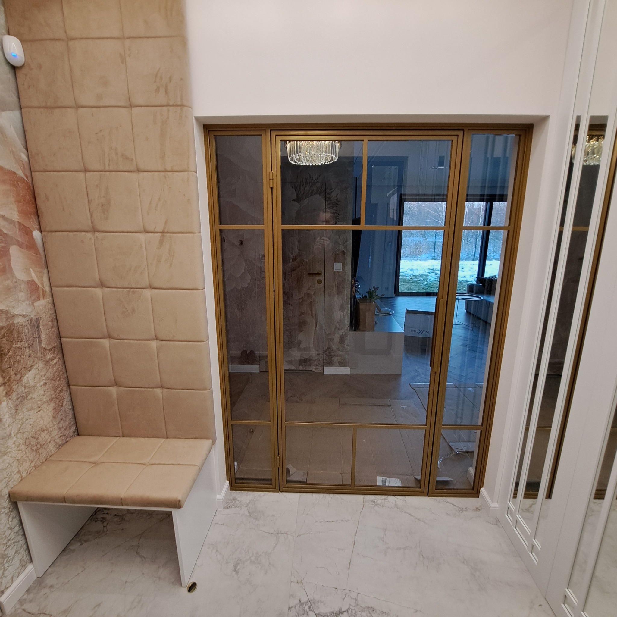 Wnętrze przedpokoju z siedziskiem obitym beżową tkaniną, ścianą z panelami tapicerowanymi i złotymi, przeszklonymi drzwiami wewnętrznymi z widokiem na dalszą część domu.