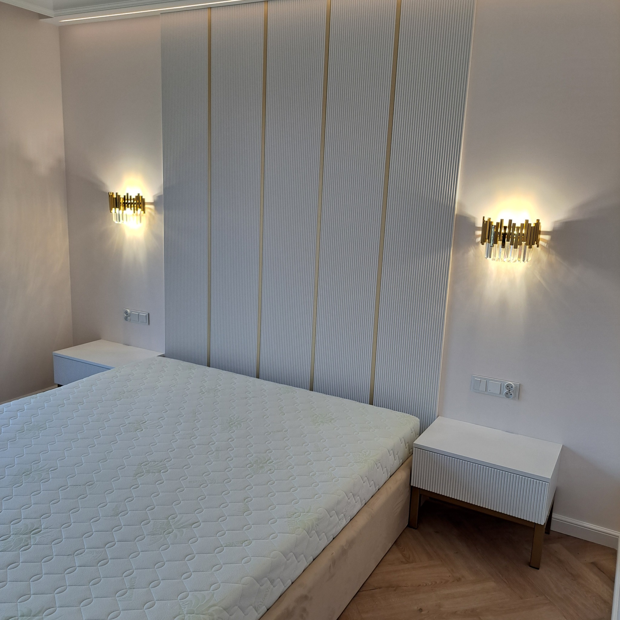 Nowoczesna sypialnia z łóżkiem z beżową ramą i materacem w geometryczne wzory, białe szafki nocne z ryflowanymi frontami i złote kinkiety na ścianie z panelami dekoracyjnymi w pionowe paski.