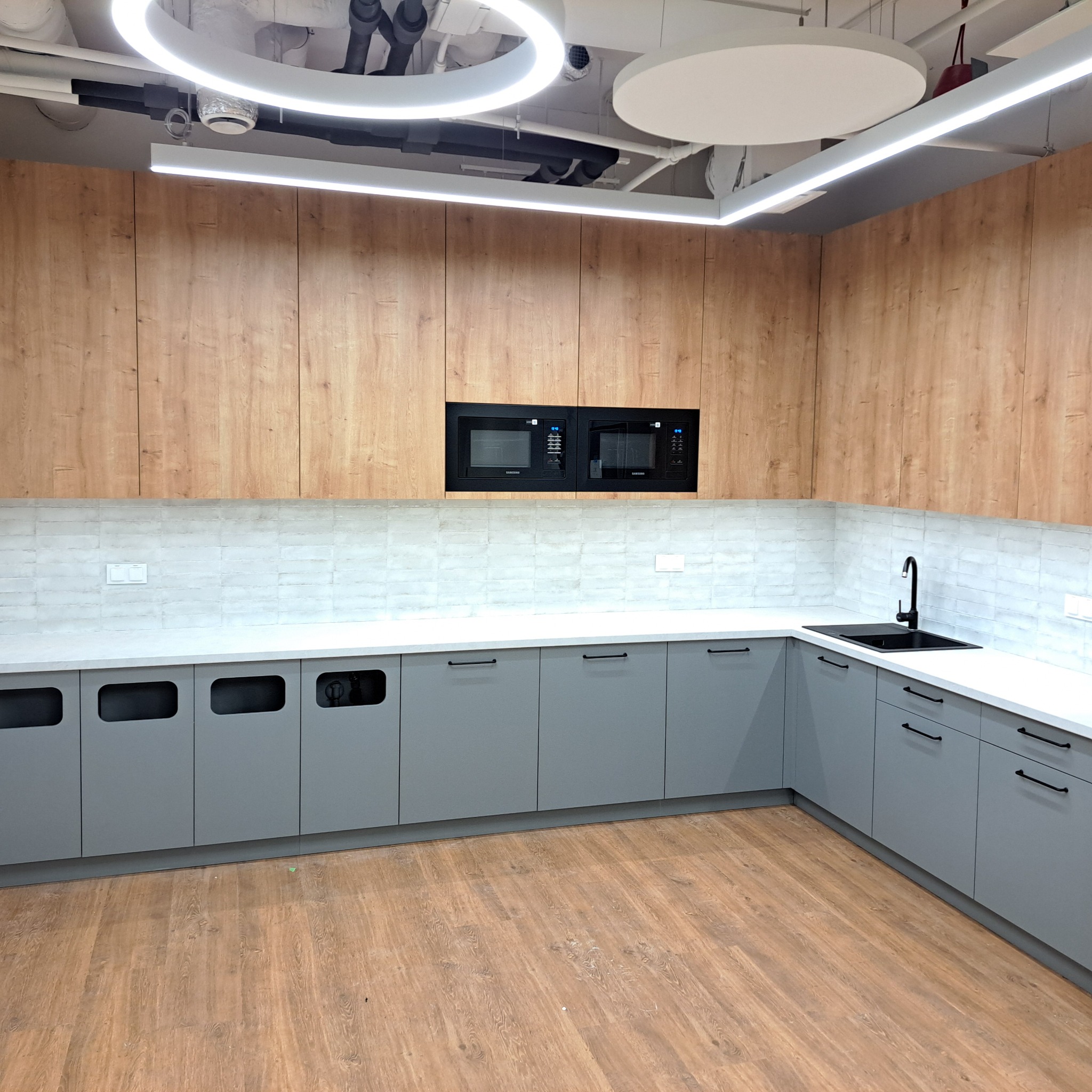 Nowoczesna kuchnia z szarymi szafkami dolnymi i drewnianymi górnymi, białym blatem, czarną armaturą i dwoma wbudowanymi mikrofalówkami Samsung, oświetlona okrągłymi lampami LED na suficie.
