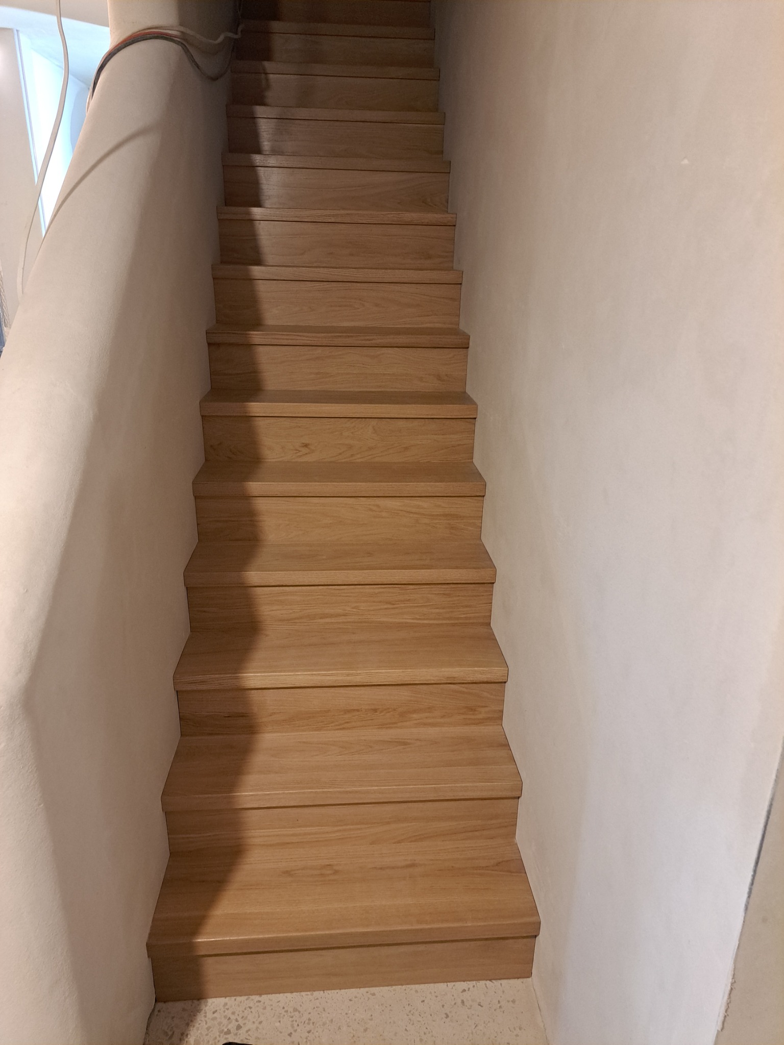 Drewniane schody o jasnym odcieniu drewna, osadzone w surowym, jasnym wnętrzu z nierównymi ścianami.