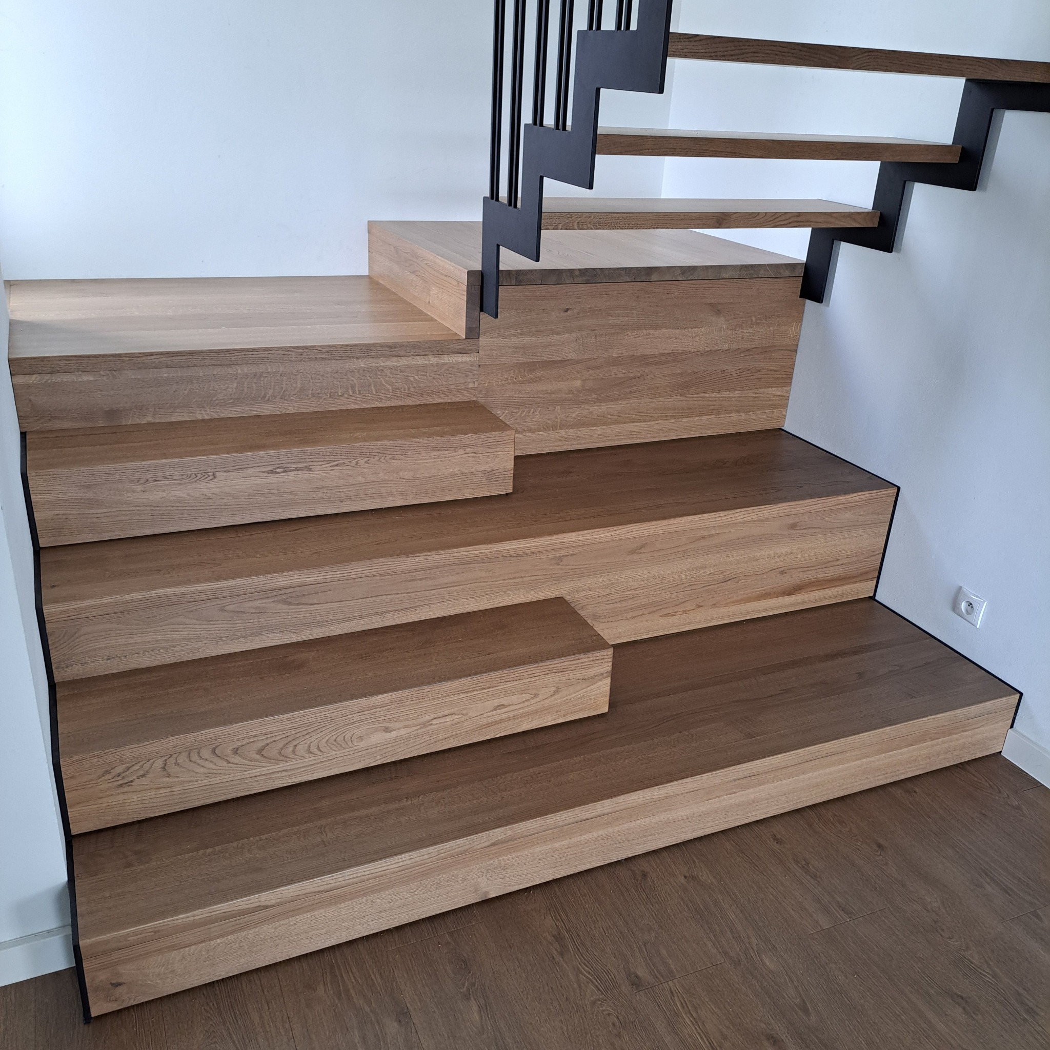 Drewniane schody z czarną, metalową balustradą o zygzakowatym wzorze na tle białej ściany z gniazdkiem elektrycznym, widoczne panele podłogowe.