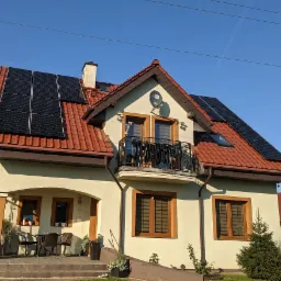 Dom jednorodzinny z czerwonym dachem i panelami fotowoltaicznymi na dachu, antena satelitarna na elewacji, balkon z ozdobną balustradą.