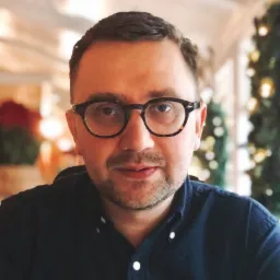 Portret mężczyzny w okularach i ciemnej koszuli na rozmytym tle z choinką, ujęcie z bliska.