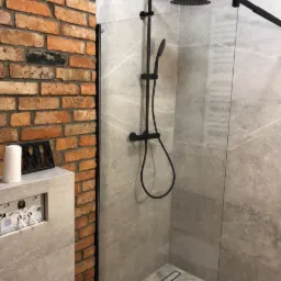 Nowoczesna kabina prysznicowa z czarną armaturą i szklaną ścianką, widoczna ceglana ściana i zabudowa WC z szarą płytką.