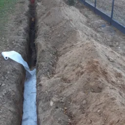 Wykop liniowy z ułożoną geowłókniną, widoczny fragment rury drenarskiej i ogrodzenie panelowe w tle.