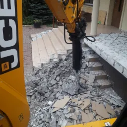 Żółta koparka JCB z młotem hydraulicznym kruszy betonowe schody przed budynkiem mieszkalnym, widok z kabiny operatora.