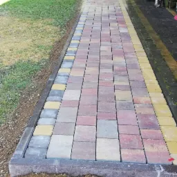 Ułożona ścieżka z wielokolorowej kostki brukowej w ogrodzie, widoczne obrzeża z ciemnej kostki, w tle trawnik i drzewa.