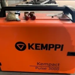 Pomarańczowa spawarka Kemppi Kempact Pulse 3000 z czarną rączką i szczotką drucianą na górze, ustawiona w warsztacie.