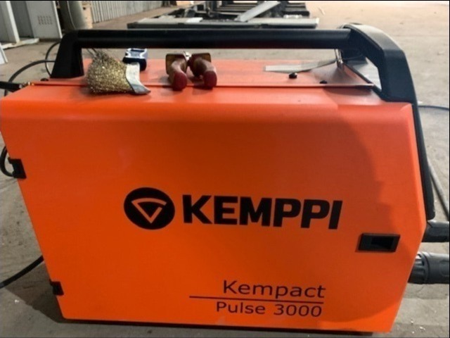 Pomarańczowa spawarka Kemppi Kempact Pulse 3000 z czarną rączką i szczotką drucianą na górze, ustawiona w warsztacie.