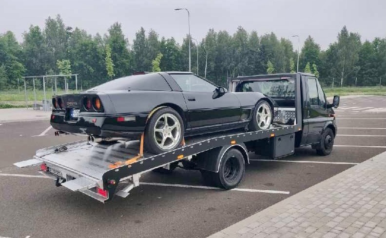 Czarna Corvette C4 na lawecie holowniczej z opuszczoną platformą najazdową na parkingu.