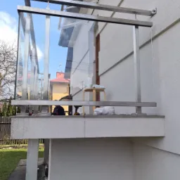 Balkon z nowoczesną, szklaną osłoną i metalową ramą, widoczne narzędzia i materiały montażowe, sugerujące trwający montaż.