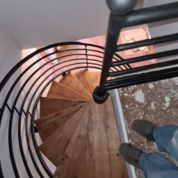 Spiralne schody z drewnianymi stopniami i czarną, metalową balustradą, widok z góry na użytkownika w jeansach i butach stojącego na górnym podeście.