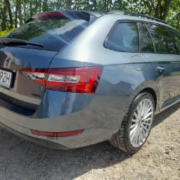 Szary samochód kombi Skoda Superb 4x4, widok z tyłu, na żwirowym podłożu, z widocznymi felgami aluminiowymi i tablicą rejestracyjną.