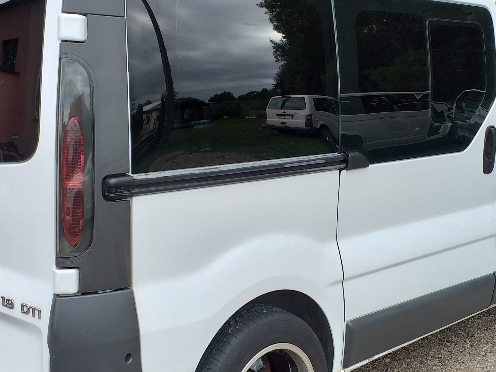 Biały bus z przyciemnianymi szybami, widoczny fragment tylnej części z reflektorem i oponą, w szybie odbija się inny bus na trawniku.