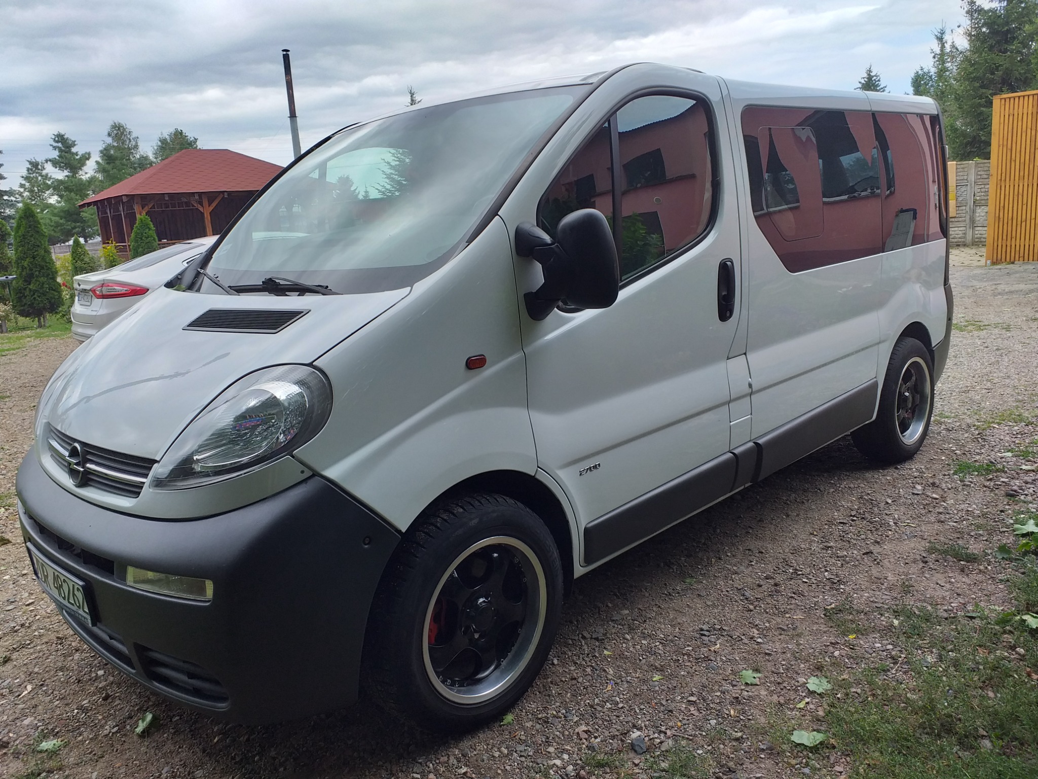 Biały Opel Vivaro z przyciemnianymi szybami i czarnymi felgami na żwirowym podjeździe, w tle altana ogrodowa i inny samochód.