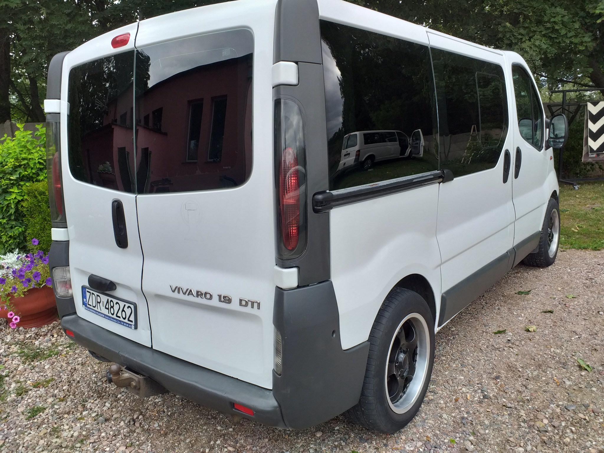 Tył białego busa Vivaro 1.9 DTI z ciemnymi szybami i hakiem holowniczym, zaparkowanego na żwirowej nawierzchni. W szybach odbija się budynek i inny bus.