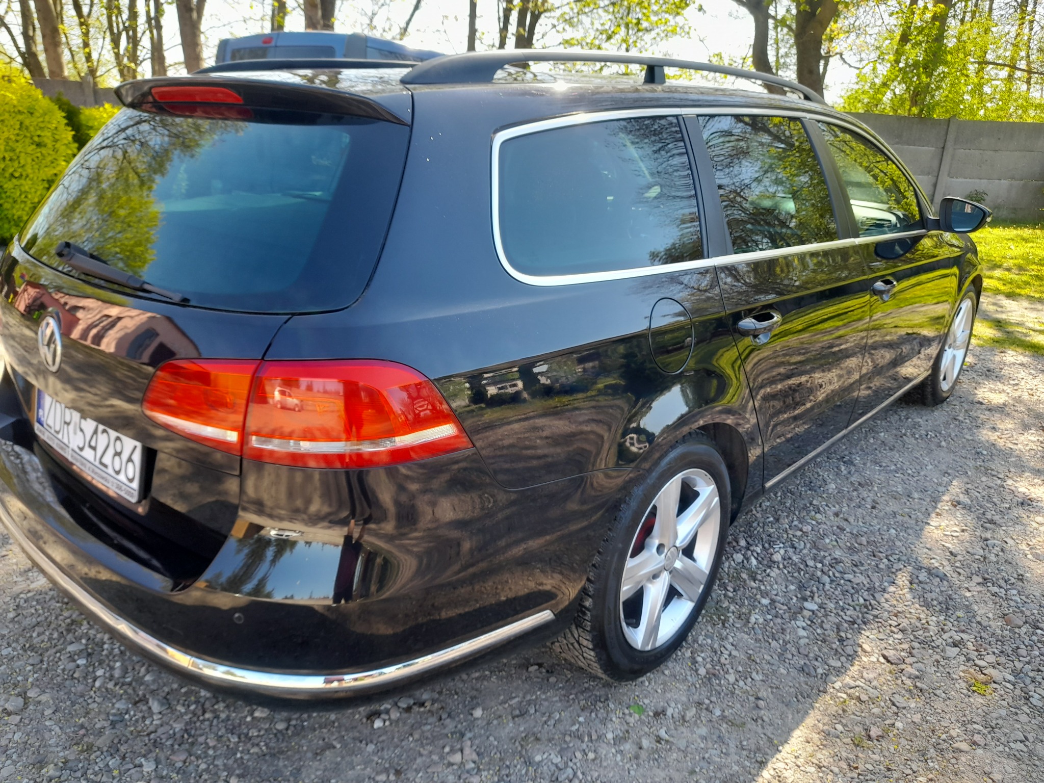 Czarny samochód kombi marki Volkswagen, widok z tyłu, zaparkowany na żwirowej nawierzchni w słoneczny dzień, z widocznymi detalami jak relingi dachowe i chromowane listwy.