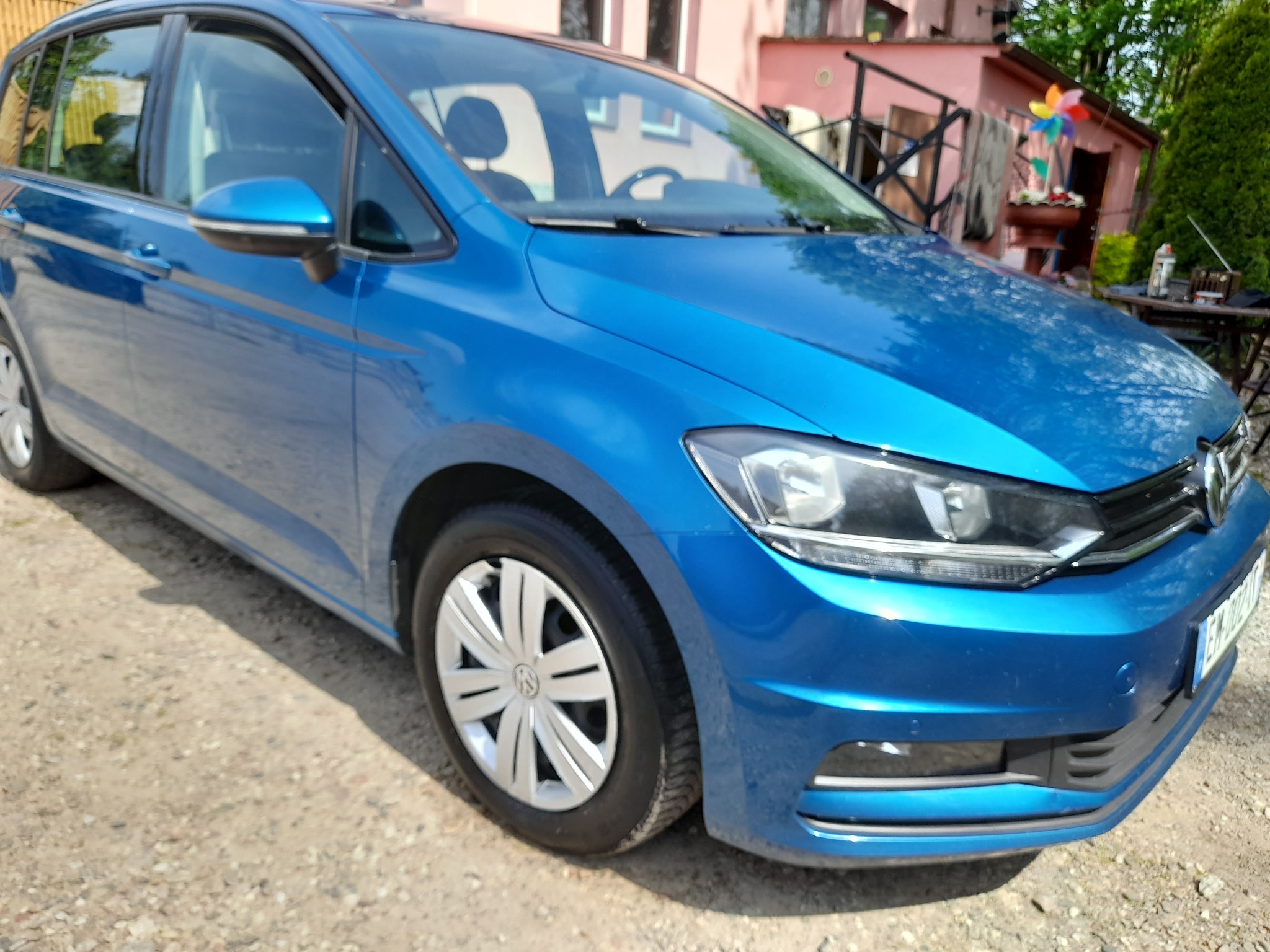 Błękitny minivan Volkswagen Touran zaparkowany na żwirowej nawierzchni, z widocznym fragmentem domu i ogrodu w tle.