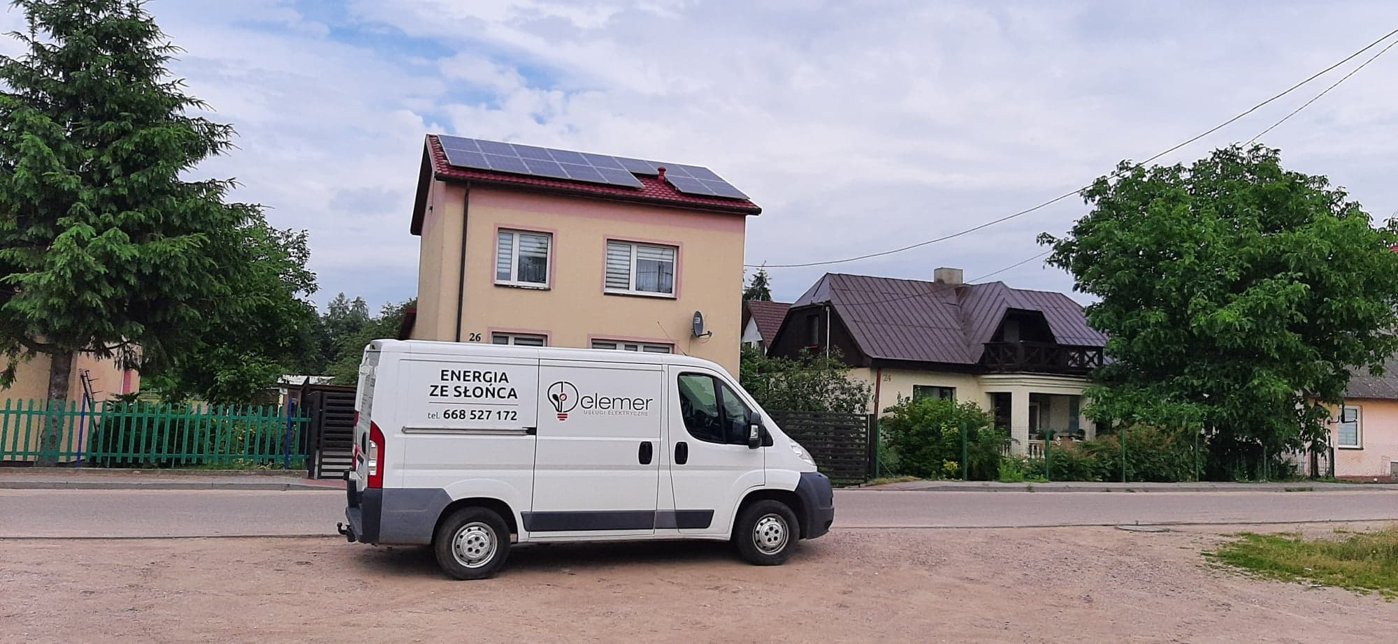 Biały samochód dostawczy z logo firmy 'Elemer' i napisem 'Energia ze Słońca' zaparkowany na tle domu z panelami słonecznymi na dachu.