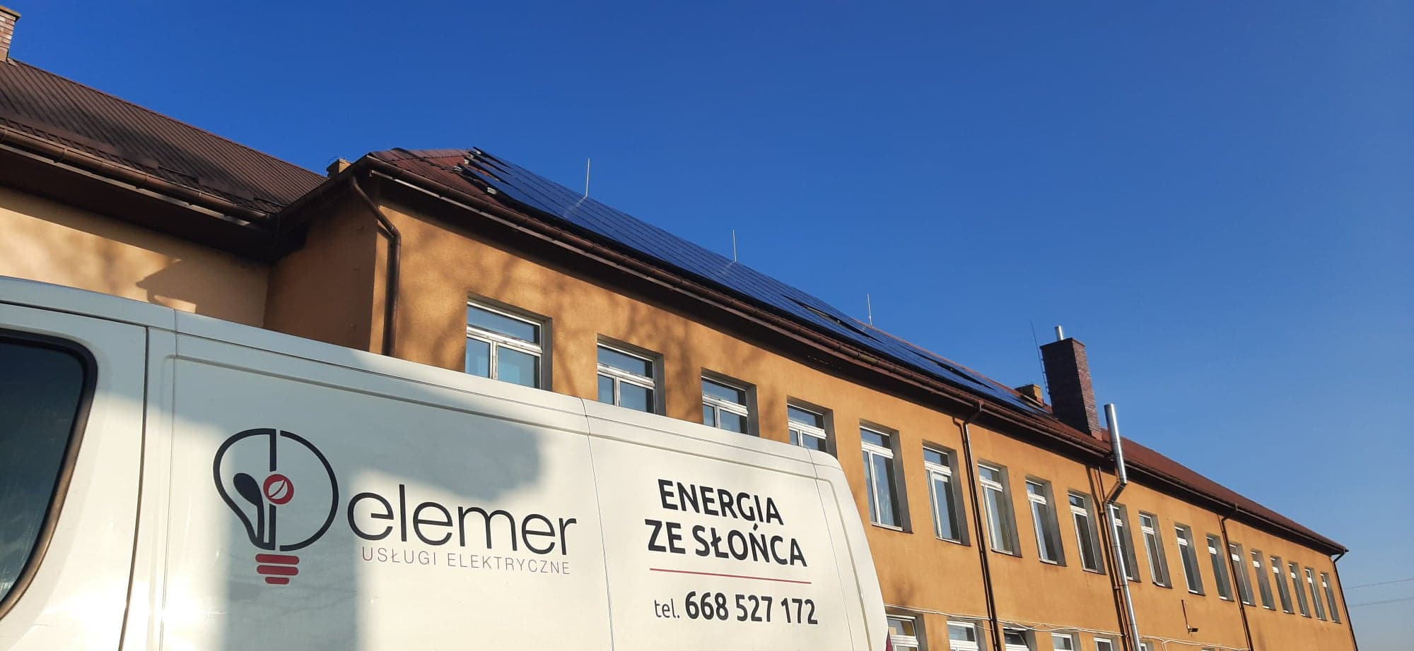 Żółty budynek z panelami fotowoltaicznymi na dachu, częściowo zasłonięty przez biały samochód dostawczy z logo firmy Elemer Usługi Elektryczne i napisem Energia ze Słońca.