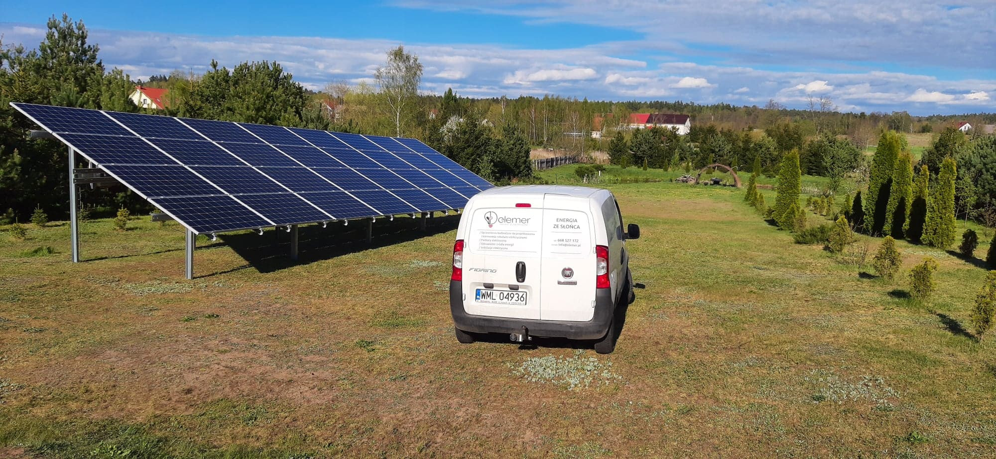 Panel słoneczny na metalowej konstrukcji, obok biały samochód dostawczy z logo firmy na tle łąki i lasu w słoneczny dzień.