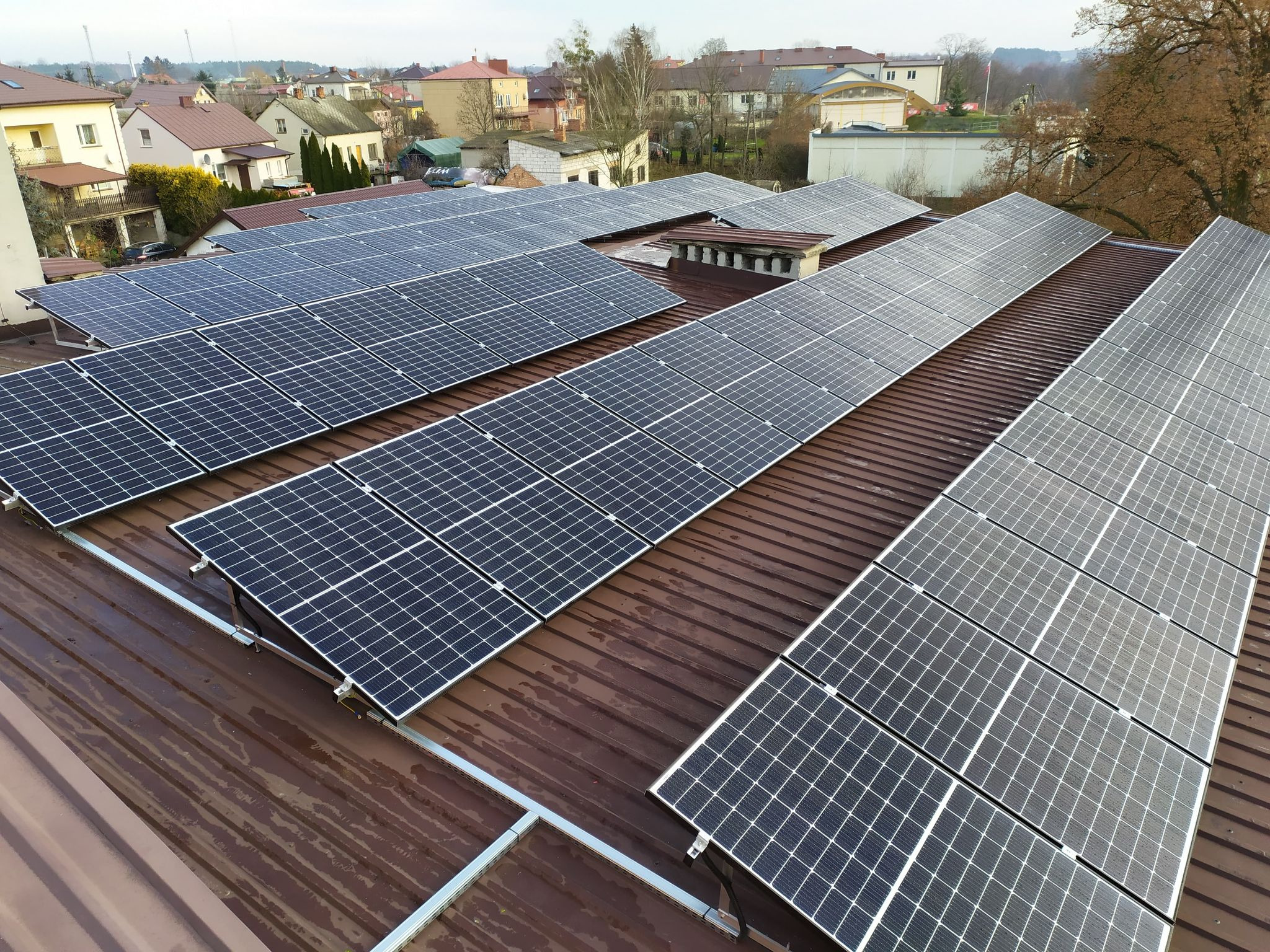 Rząd paneli słonecznych na brązowym, metalowym dachu budynku mieszkalnego, widok z góry, w tle zabudowa miejska