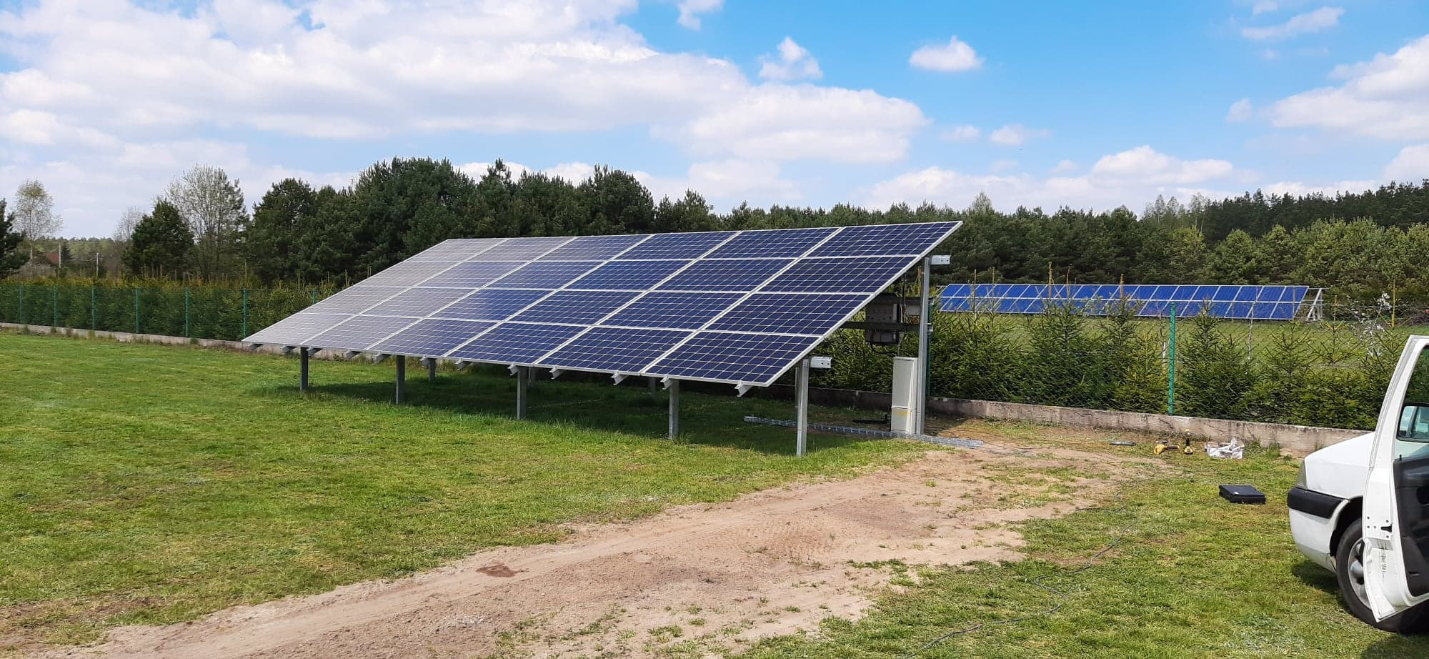 Instalacja fotowoltaiczna na trawiastym terenie z widocznymi panelami, konstrukcją wsporczą i skrzynką przyłączeniową, w tle las i błękitne niebo z chmurami, częściowo widoczny samochód dostawczy.