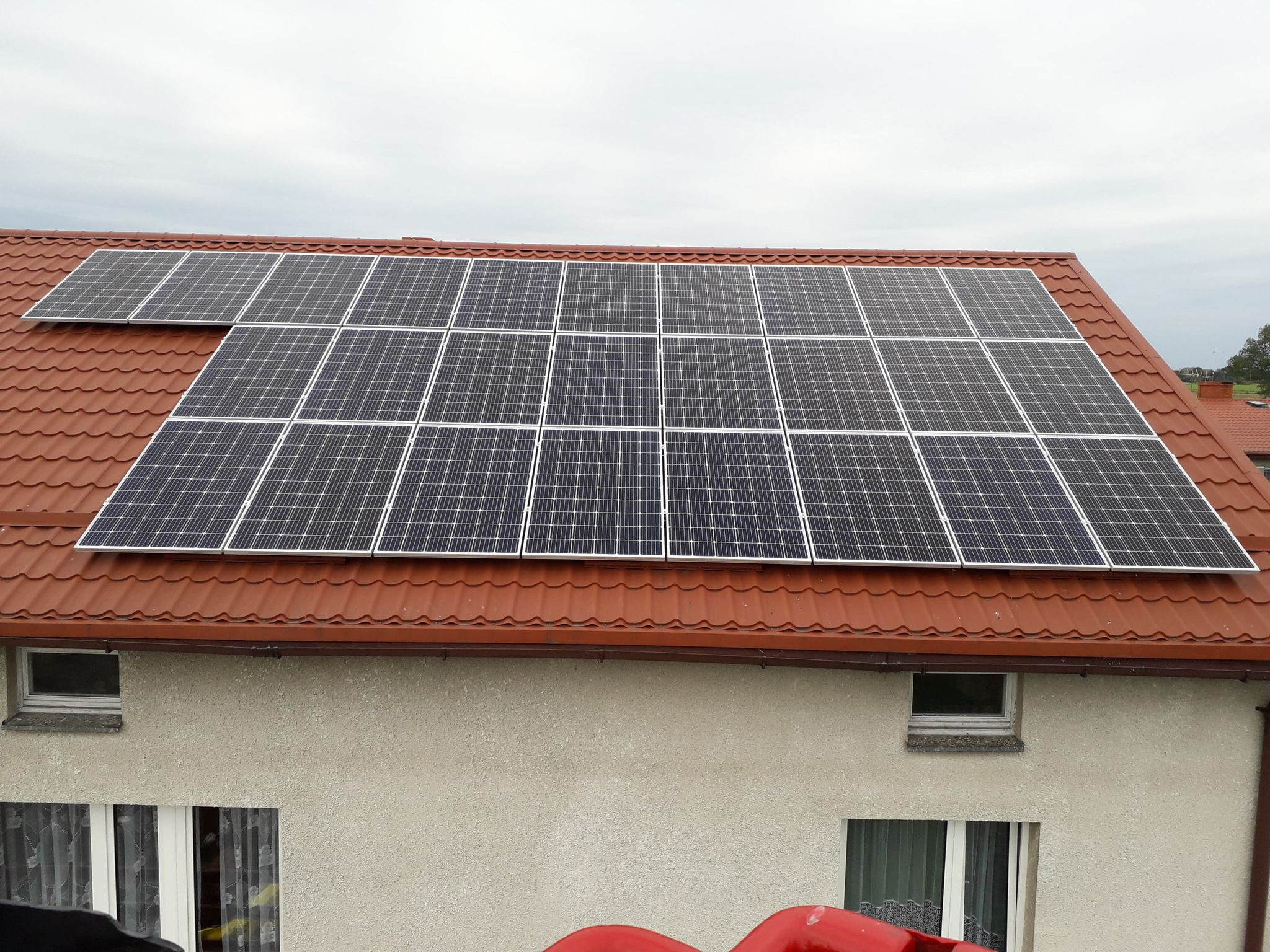 Panele słoneczne zamontowane na dachu z czerwonej dachówki, widok z góry na instalację z jasną elewacją budynku w tle.