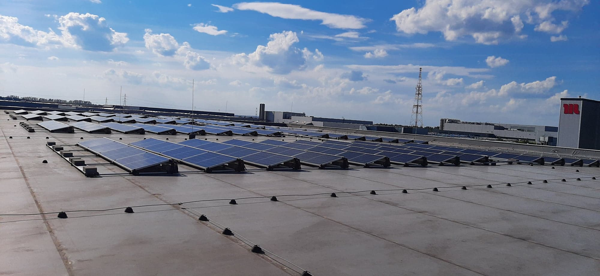Rząd paneli słonecznych na płaskim dachu budynku przemysłowego w słoneczny dzień z błękitnym niebem i białymi chmurami w tle.