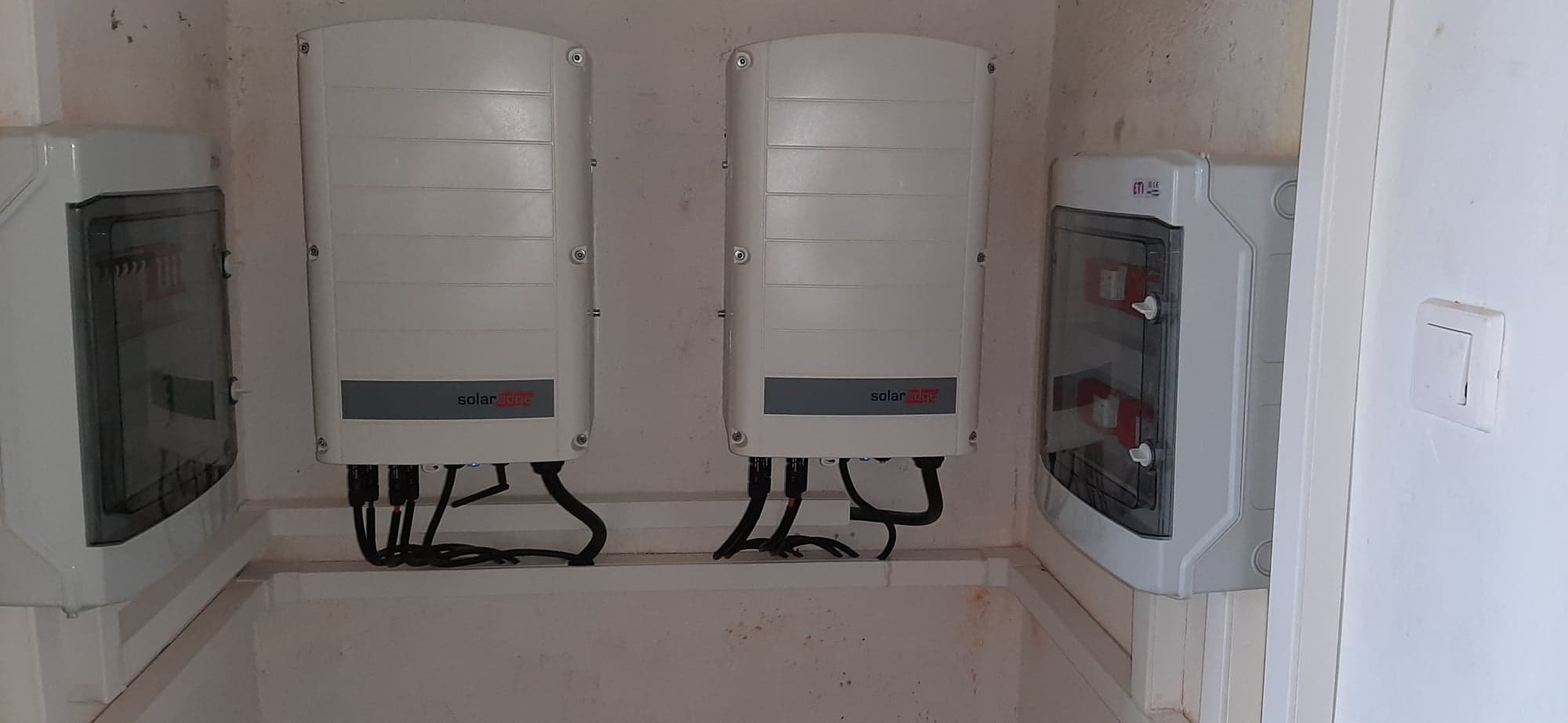 Dwa falowniki SolarEdge zamontowane obok siebie na ścianie z widocznymi przewodami, obok skrzynka elektryczna z widocznymi bezpiecznikami oraz włącznik światła.