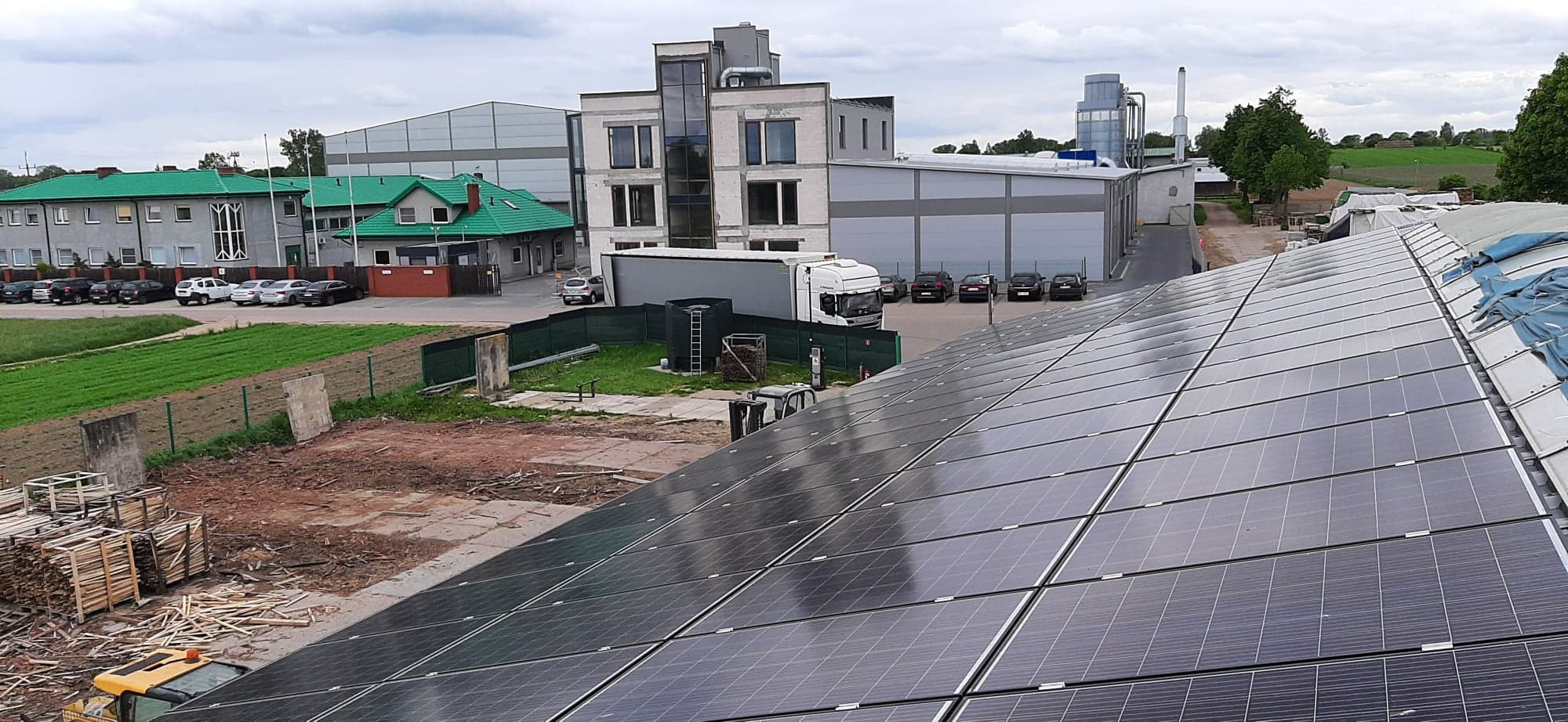 Rozległa instalacja fotowoltaiczna na dachu budynku przemysłowego, z widocznymi panelami słonecznymi, częściowo zakrytymi plandeką, w tle budynki biurowe i przemysłowe, zaparkowane samochody...