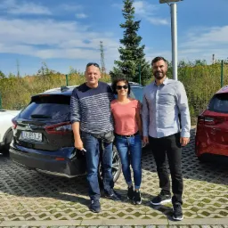 Trzy osoby stojące obok zaparkowanych samochodów na betonowym parkingu w słoneczny dzień. W tle widoczna zieleń i słup oświetleniowy z panelem solarnym.