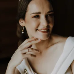 Portret uśmiechniętej panny młodej w białej sukni z perłową opaską, złotymi kolczykami i pierścionkiem zaręczynowym, delikatnie dotykającej podbródka.