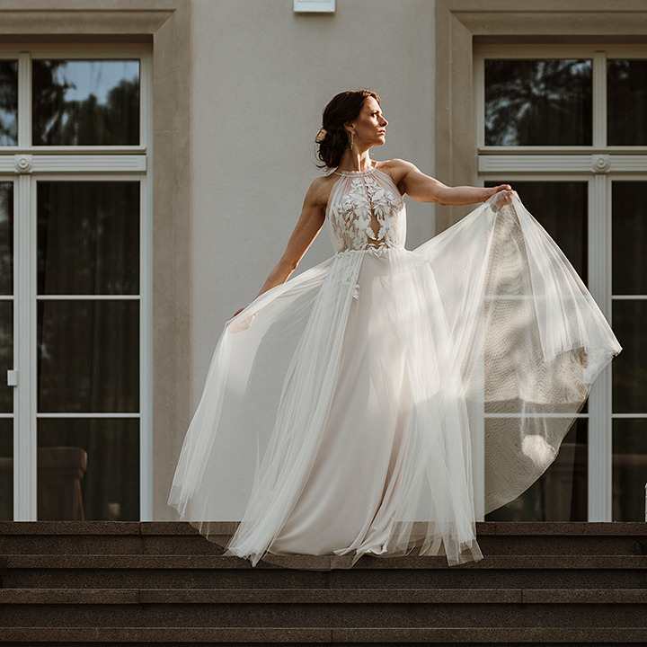 Panna młoda w zwiewnej sukni z koronkowym gorsetem, pozuje na schodach klasycystycznego budynku z białymi oknami, spoglądając w bok.