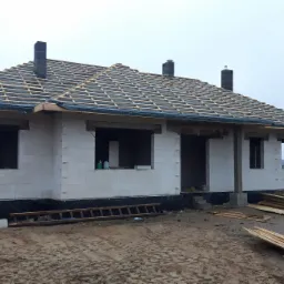 Surowy stan domu jednorodzinnego z białych bloczków, widoczna konstrukcja dachu z czarną membraną, kominy, teren budowy z drewnem i pustakami.