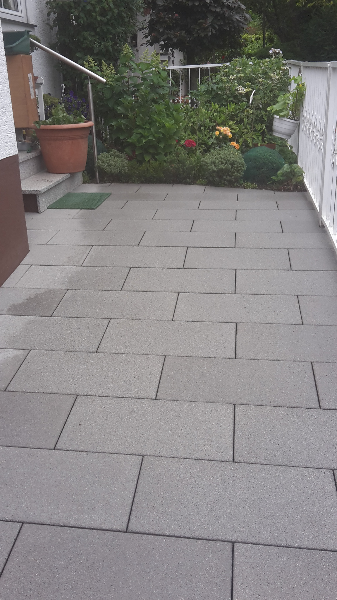 Ułożona jasnoszara kostka brukowa w Jastrowiu, prowadząca od schodów do białego ogrodzenia, z widoczną zielenią roślin w tle.