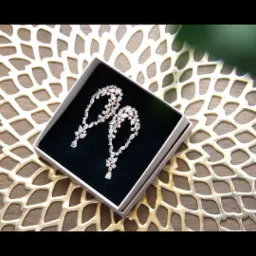 Dwa błyszczące, wiszące kolczyki z kryształkami w białym pudełku na złotej, ażurowej podkładce, z delikatnym rozmyciem rośliny w tle.