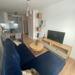 Akwadrat Deweloper Sp. z o.o. - Nowoczesny salon z granatową sofą narożną, okrągłym stolikiem kawowym z kwiatem i drewnianą szafką RTV pod telewizorem, uzupełniony wiszącymi półkami i lampami.