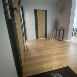 Akwadrat Deweloper Sp. z o.o. - Nowoczesne wnętrze z drewnianą podłogą, czarnymi futrynami drzwiowymi i dekoracyjnym dywanem z motywem roślinnym w rogu.