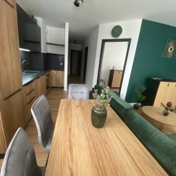 Akwadrat Deweloper Sp. z o.o. - Nowoczesne wnętrze: drewniany stół z szarymi krzesłami, kuchnia z czarnymi frontami i drewnianymi szafkami, zielona ściana z dekoracjami, widok na korytarz.