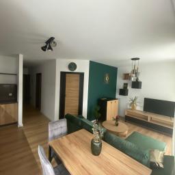 Akwadrat Deweloper Sp. z o.o. - Jasne wnętrze salonu z drewnianą podłogą, zieloną ścianą akcentującą, drewnianym stołem i szarymi krzesłami, widok na korytarz z drewnianymi drzwiami.