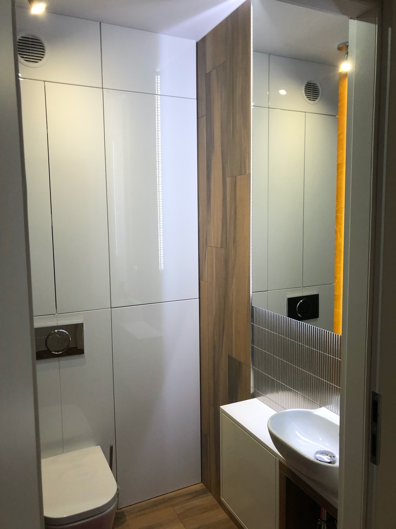 Nowoczesna łazienka z białą zabudową meblową, wiszącą miską WC z chromowanym przyciskiem spłukiwania, umywalką nablatową i dekoracyjną ścianką z płytek imitujących drewno