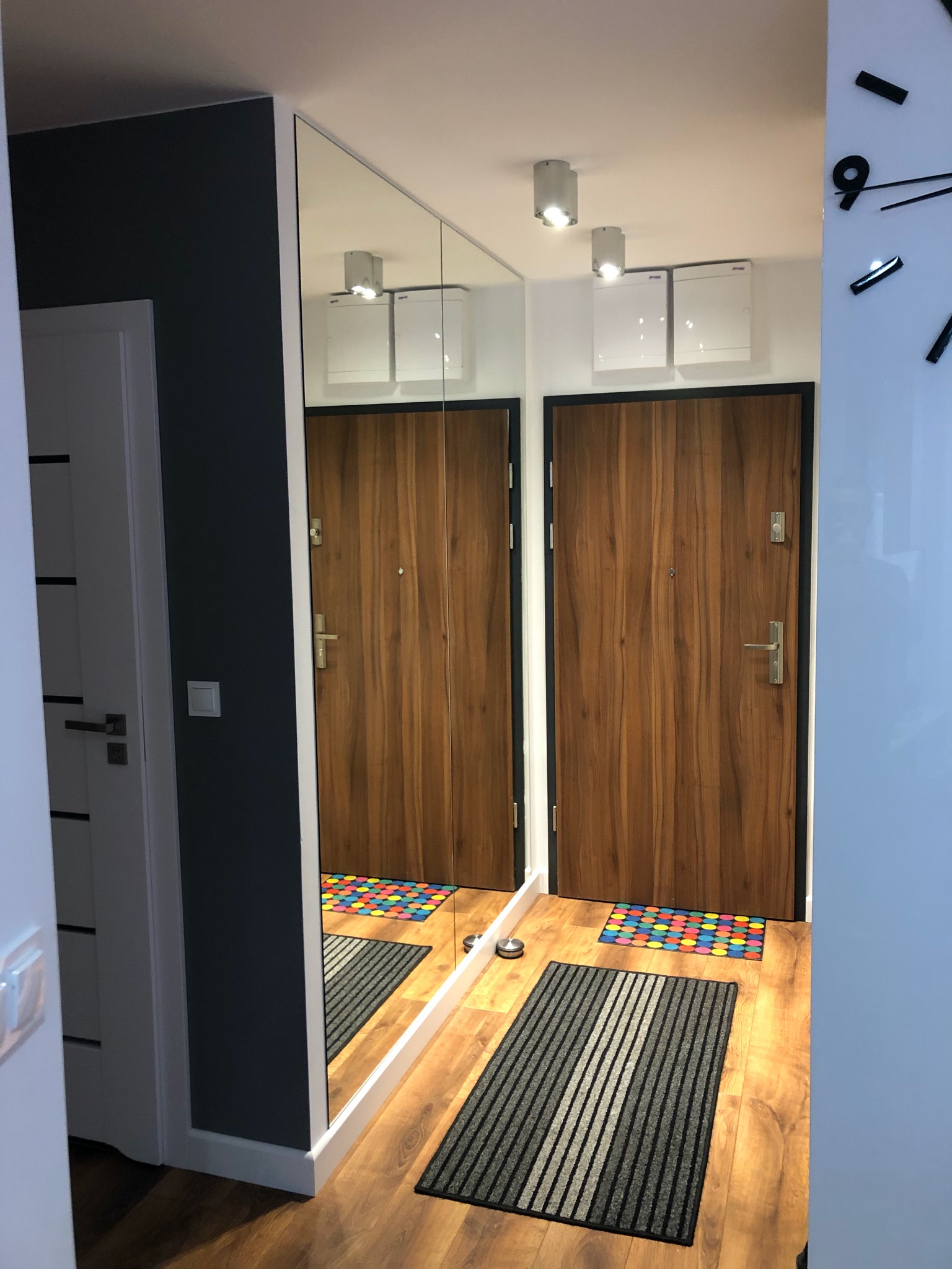 Nowoczesny przedpokój z drewnianymi drzwiami wejściowymi, lustrem w zabudowie i parkietem, uzupełniony o szarą wycieraczkę w paski i kolorową wycieraczkę pod drzwiami.